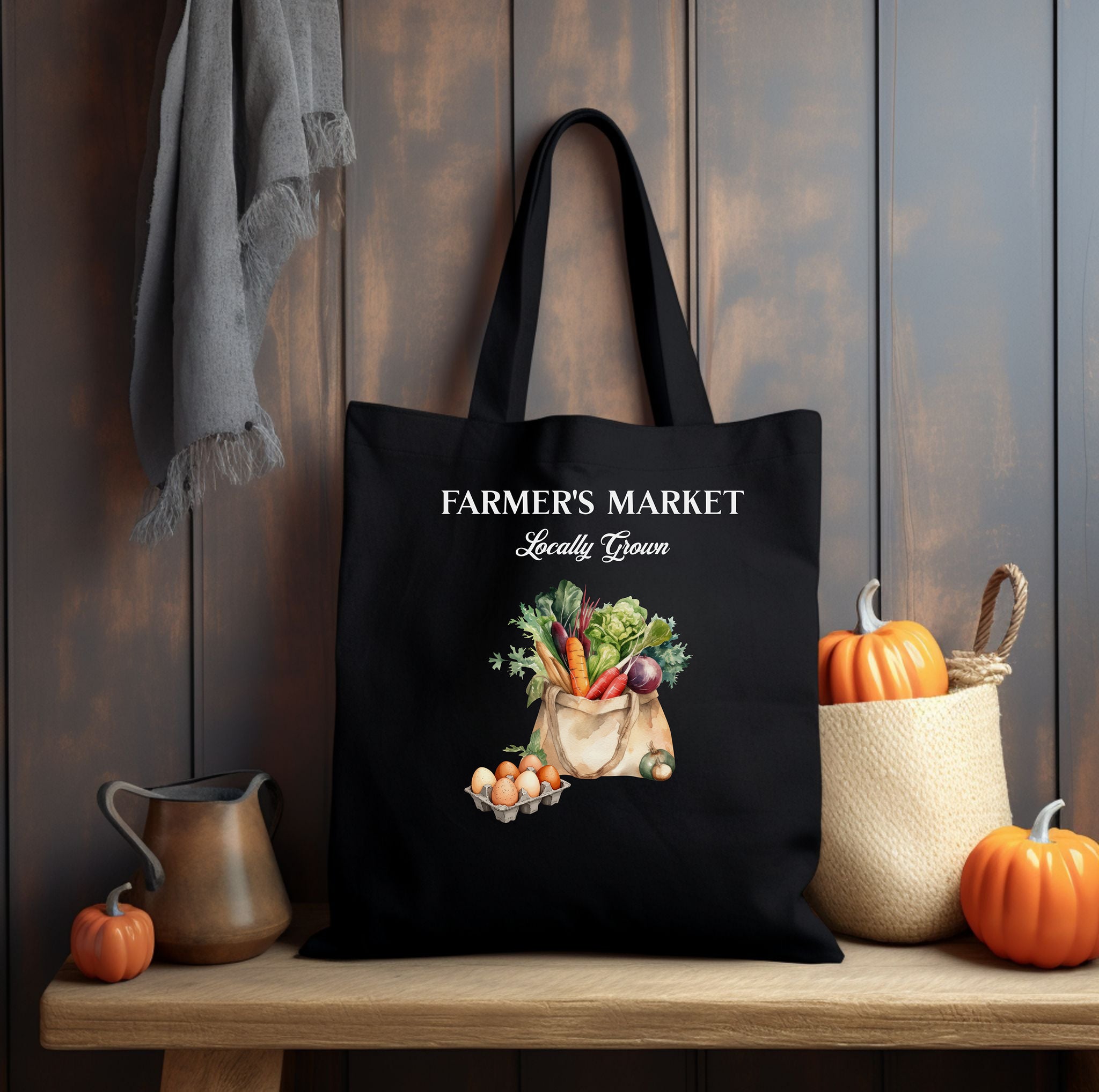 A Blackl Tote bag with 'Farmer's Market Locally Grown' white text and vegetable in a tote bag illustration with carton of eggs next to it sitting on a table with pumpkins and autumn decor around it.