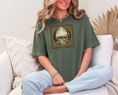 Hemp color “Fellowship” T-shirt featuring a fantasy-inspired design of five cloaked figures walking along a stone path toward distant mountains. The scene is framed by intricate Celtic-style borders with bare trees arching overhead, creating an adventurous and mythical atmosphere. Perfect for fans of epic journeys, fantasy worlds, and storytelling-inspired apparel.