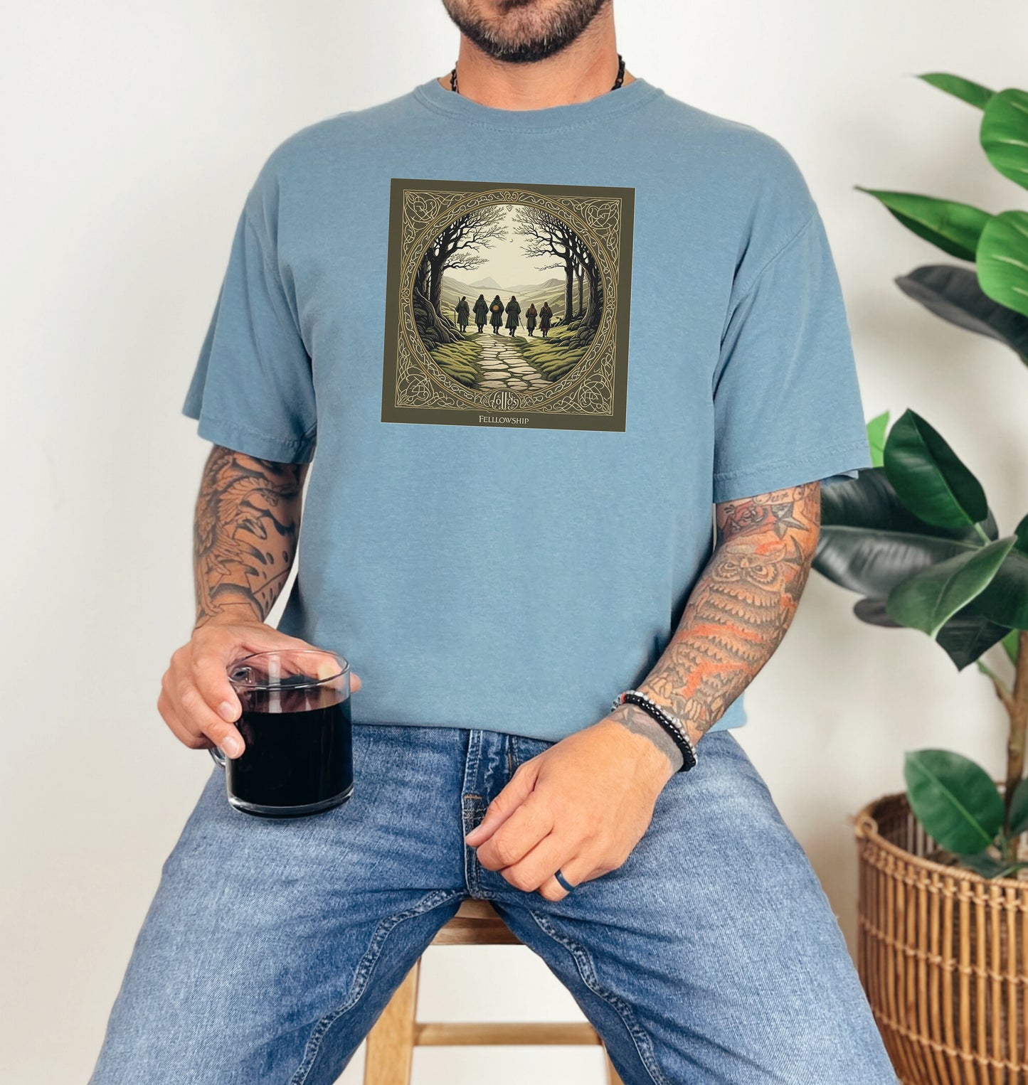 Ice Blue “Fellowship” T-shirt featuring a fantasy-inspired design of five cloaked figures walking along a stone path toward distant mountains. The scene is framed by intricate Celtic-style borders with bare trees arching overhead, creating an adventurous and mythical atmosphere. Perfect for fans of epic journeys, fantasy worlds, and storytelling-inspired apparel.