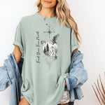 Person wearing a bay sage green T-shirt featuring a “Find Your True North” design with a vertical compass, full moon, pine trees, mountains, soaring eagle, and howling wolf in detailed black and white sketch style. The design blends celestial and wilderness elements, symbolizing adventure, guidance, and a connection to nature. Perfect for outdoor lovers, travelers, and free spirits seeking inspiration and direction.