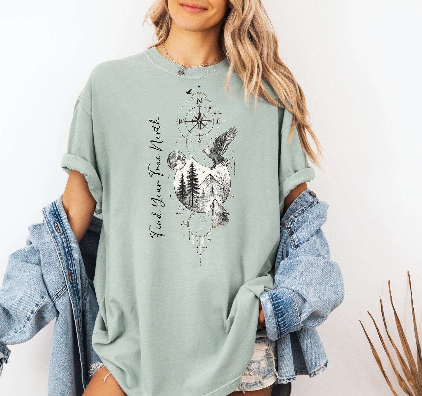 Person wearing a bay sage green T-shirt featuring a “Find Your True North” design with a vertical compass, full moon, pine trees, mountains, soaring eagle, and howling wolf in detailed black and white sketch style. The design blends celestial and wilderness elements, symbolizing adventure, guidance, and a connection to nature. Perfect for outdoor lovers, travelers, and free spirits seeking inspiration and direction.
