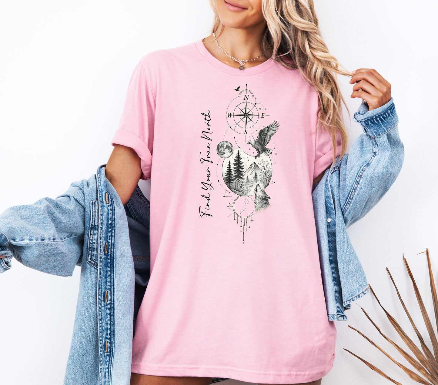 Person wearing a blossom pink T-shirt featuring a “Find Your True North” design with a vertical compass, full moon, pine trees, mountains, soaring eagle, and howling wolf in detailed black and white sketch style. The design blends celestial and wilderness elements, symbolizing adventure, guidance, and a connection to nature. Perfect for outdoor lovers, travelers, and free spirits seeking inspiration and direction.