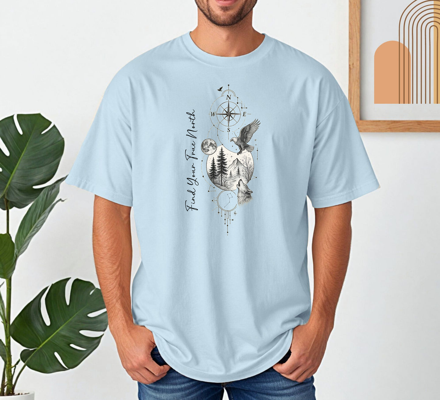 Person wearing a chambray light blue T-shirt featuring a “Find Your True North” design with a vertical compass, full moon, pine trees, mountains, soaring eagle, and howling wolf in detailed black and white sketch style. The design blends celestial and wilderness elements, symbolizing adventure, guidance, and a connection to nature. Perfect for outdoor lovers, travelers, and free spirits seeking inspiration and direction.