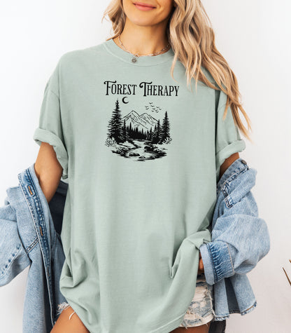 Person wearing a bay light green t-shirt featuring a black “Forest Therapy” design with a peaceful mountain scene, pine trees, a flowing river, birds, and a crescent moon. The perfect tee for nature lovers, hikers, and outdoor enthusiasts seeking serenity and scenic vibes.