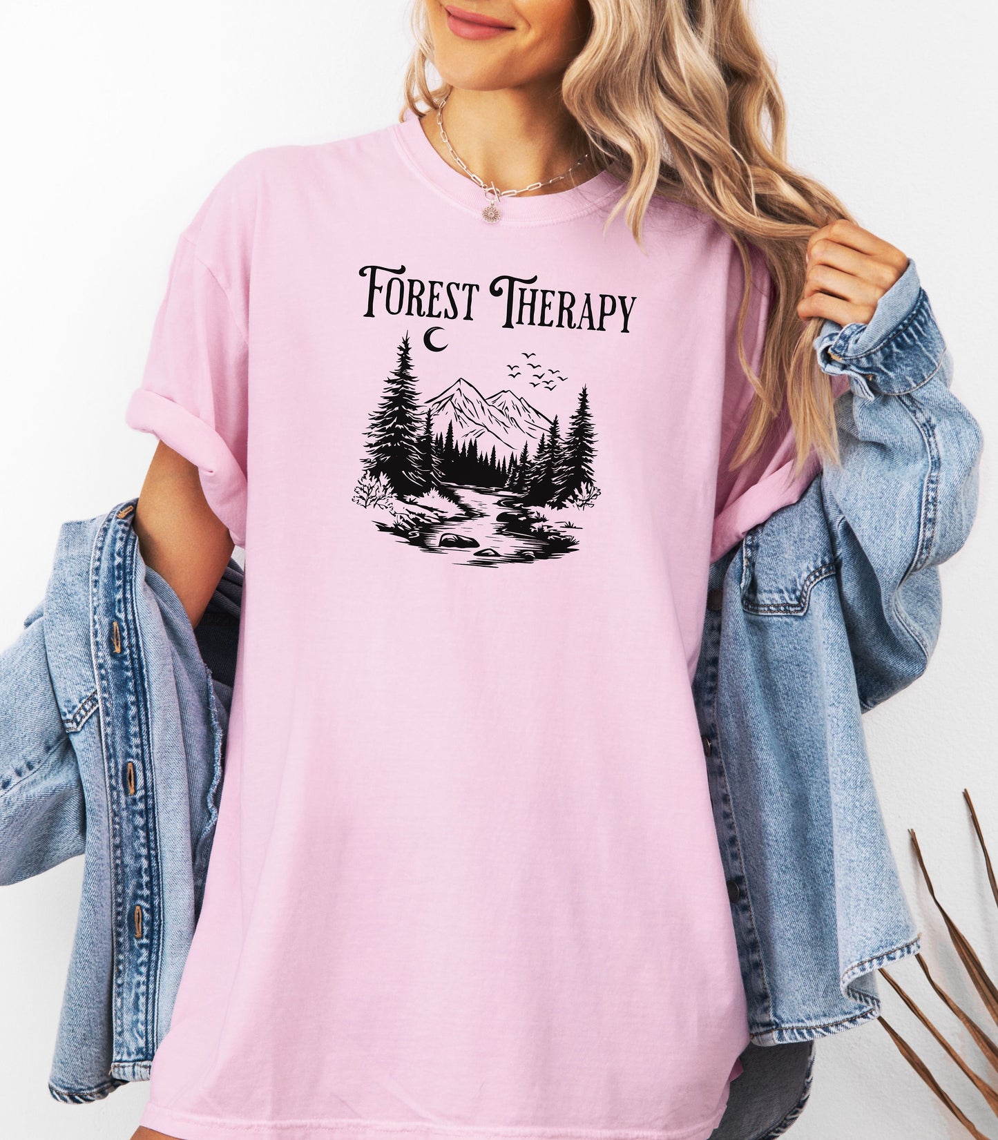 Person wearing a Blossom pink t-shirt featuring a black “Forest Therapy” design with a peaceful mountain scene, pine trees, a flowing river, birds, and a crescent moon. The perfect tee for nature lovers, hikers, and outdoor enthusiasts seeking serenity and scenic vibes.