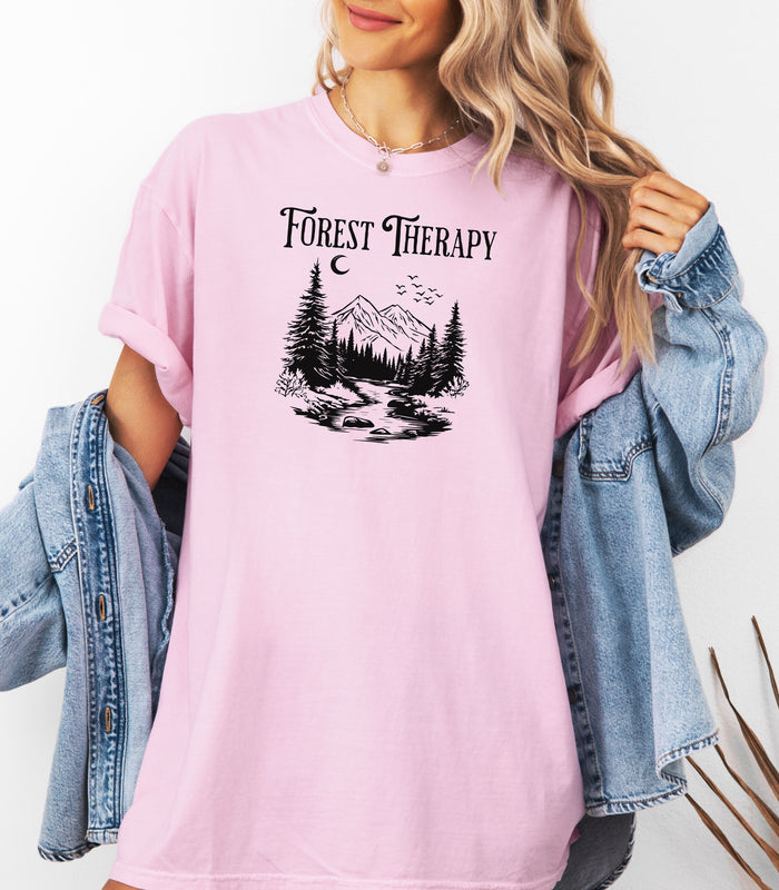 Person wearing a Blossom pink t-shirt featuring a black “Forest Therapy” design with a peaceful mountain scene, pine trees, a flowing river, birds, and a crescent moon. The perfect tee for nature lovers, hikers, and outdoor enthusiasts seeking serenity and scenic vibes.