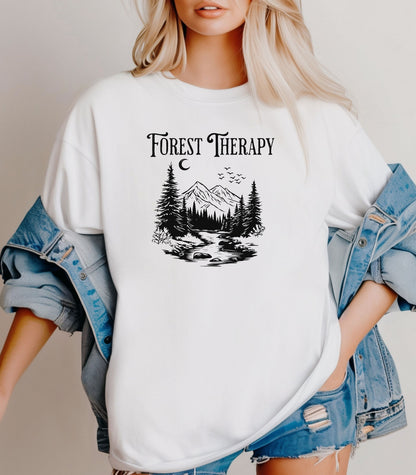 Person wearing a White t-shirt featuring a black “Forest Therapy” design with a peaceful mountain scene, pine trees, a flowing river, birds, and a crescent moon. The perfect tee for nature lovers, hikers, and outdoor enthusiasts seeking serenity and scenic vibes.