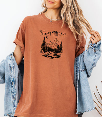 Person wearing a yam t-shirt featuring a black “Forest Therapy” design with a peaceful mountain scene, pine trees, a flowing river, birds, and a crescent moon. The perfect tee for nature lovers, hikers, and outdoor enthusiasts seeking serenity and scenic vibes.
