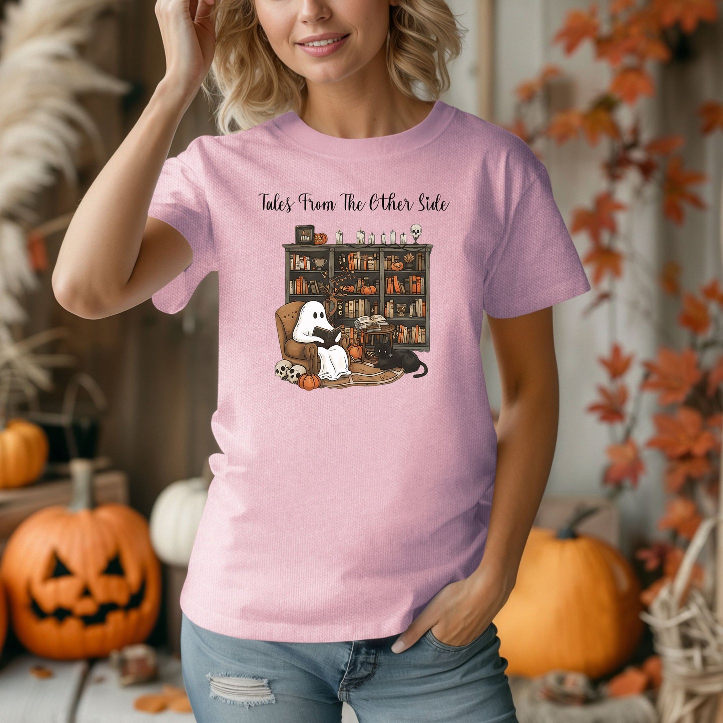 Person wearing a blossom pink t-shirt featuring a cozy Halloween-themed design with a ghost reading a book in an armchair, a black cat beside it, a skull, pumpkins, and a bookshelf filled with spooky books and potion bottles. The text reads "Tales From The Other Side," creating a whimsical and gothic vibe perfect for book lovers and Halloween fans.