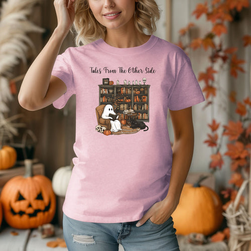 Person wearing a blossom pink t-shirt featuring a cozy Halloween-themed design with a ghost reading a book in an armchair, a black cat beside it, a skull, pumpkins, and a bookshelf filled with spooky books and potion bottles. The text reads "Tales From The Other Side," creating a whimsical and gothic vibe perfect for book lovers and Halloween fans.
