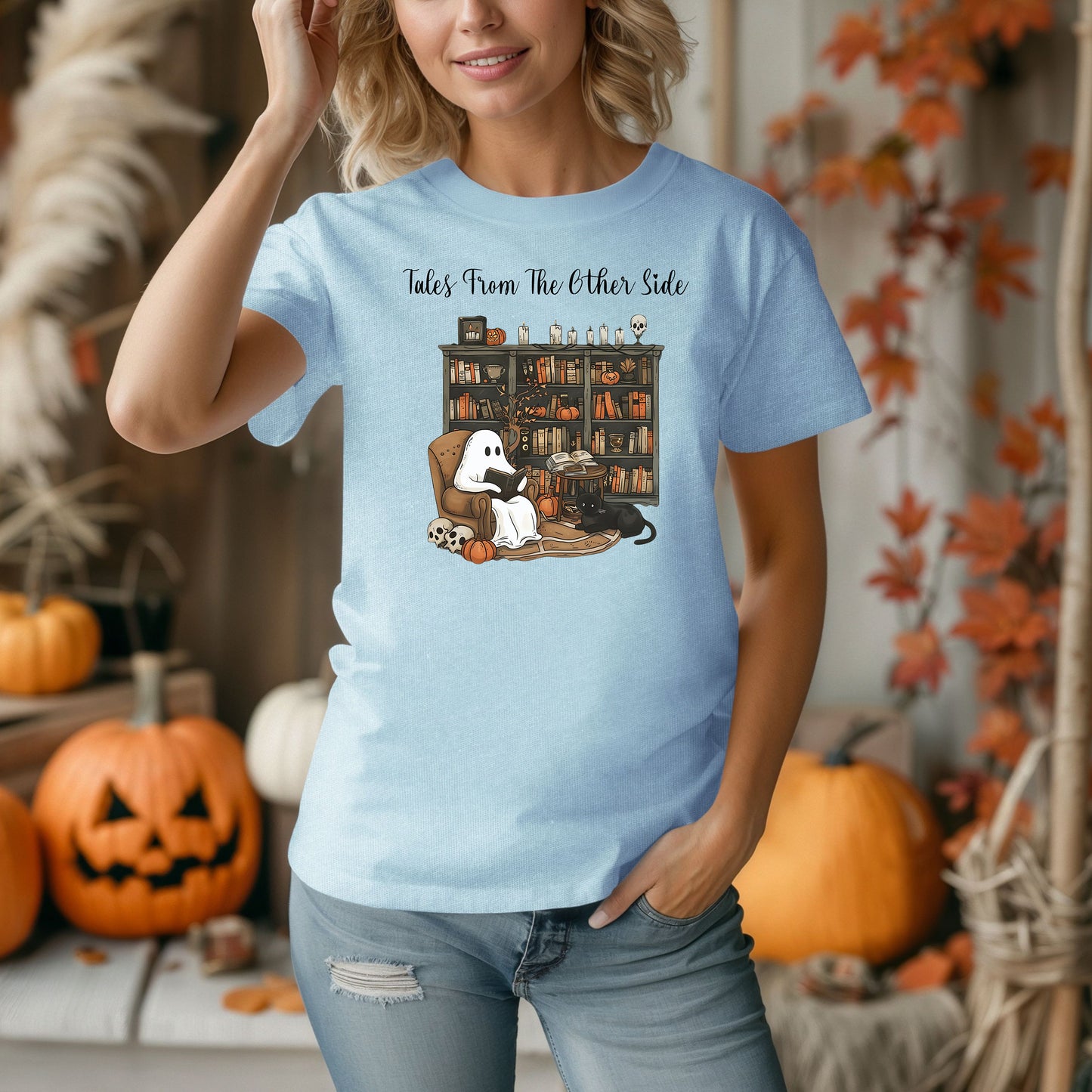 Person wearing a Chambray light blue t-shirt featuring a cozy Halloween-themed design with a ghost reading a book in an armchair, a black cat beside it, a skull, pumpkins, and a bookshelf filled with spooky books and potion bottles. The text reads "Tales From The Other Side," creating a whimsical and gothic vibe perfect for book lovers and Halloween fans.