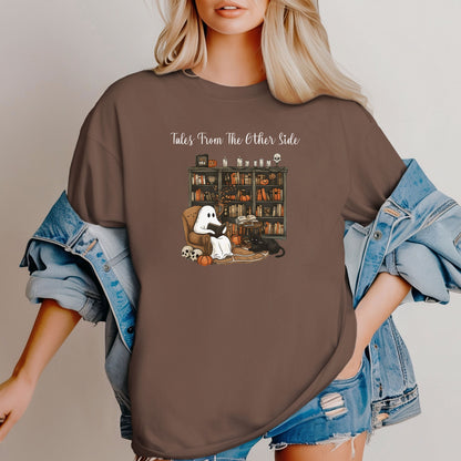 Person wearing an Espresso t-shirt featuring a cozy Halloween-themed design with a ghost reading a book in an armchair, a black cat beside it, a skull, pumpkins, and a bookshelf filled with spooky books and potion bottles. The text reads "Tales From The Other Side," creating a whimsical and gothic vibe perfect for book lovers and Halloween fans.