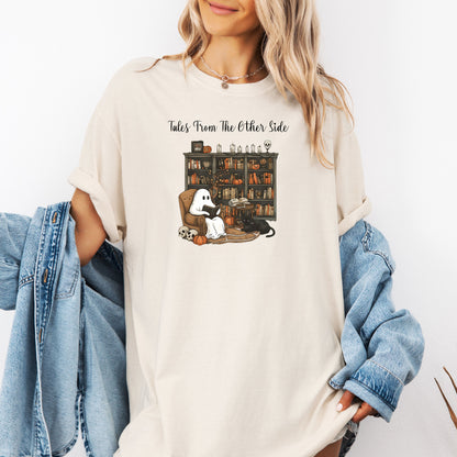 Person wearing an Ivory t-shirt featuring a cozy Halloween-themed design with a ghost reading a book in an armchair, a black cat beside it, a skull, pumpkins, and a bookshelf filled with spooky books and potion bottles. The text reads "Tales From The Other Side," creating a whimsical and gothic vibe perfect for book lovers and Halloween fans.