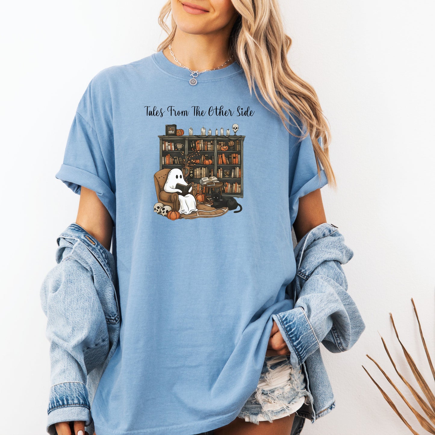Person wearing a Washed Denim t-shirt featuring a cozy Halloween-themed design with a ghost reading a book in an armchair, a black cat beside it, a skull, pumpkins, and a bookshelf filled with spooky books and potion bottles. The text reads "Tales From The Other Side," creating a whimsical and gothic vibe perfect for book lovers and Halloween fans.