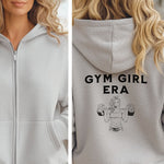 Ash Gildan hoodie featuring bold black text “GYM GIRL ERA” on the back, above a line art illustration of a muscular woman confidently lifting heavy barbells. The empowering fitness design celebrates strength, dedication, and gym lifestyle—perfect for active women embracing their fitness journey.