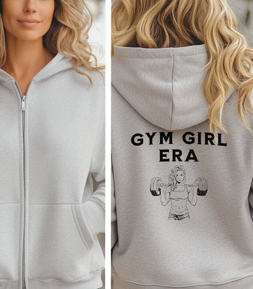 Ash Gildan hoodie featuring bold black text “GYM GIRL ERA” on the back, above a line art illustration of a muscular woman confidently lifting heavy barbells. The empowering fitness design celebrates strength, dedication, and gym lifestyle—perfect for active women embracing their fitness journey.