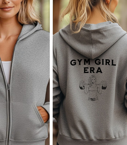 Sport Grey Gildan hoodie featuring bold black text “GYM GIRL ERA” on the back, above a line art illustration of a muscular woman confidently lifting heavy barbells. The empowering fitness design celebrates strength, dedication, and gym lifestyle—perfect for active women embracing their fitness journey.