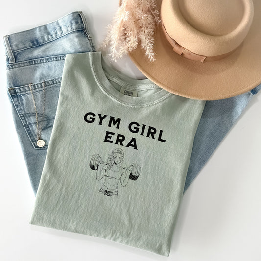 A bay T-shirt with the bold black text “Gym Girl Era” above a line art illustration of a muscular woman lifting dumbbells. The fitness-inspired design celebrates strength, empowerment, and dedication to the gym lifestyle. Paired with denim shorts and a jean jacket, the look is casual yet powerful.