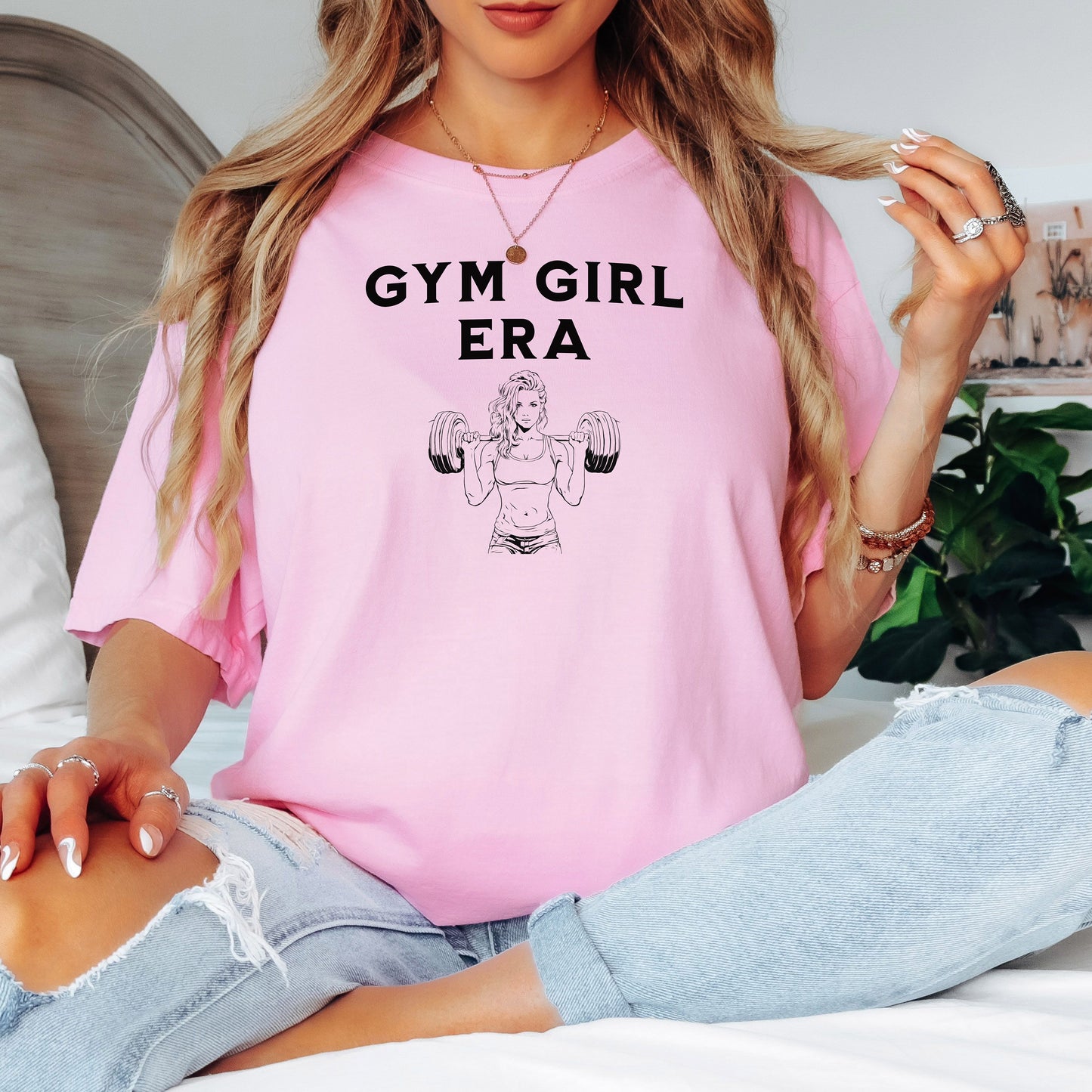 A blossom pink T-shirt with the bold black text “Gym Girl Era” above a line art illustration of a muscular woman lifting dumbbells. The fitness-inspired design celebrates strength, empowerment, and dedication to the gym lifestyle. Paired with denim shorts and a jean jacket, the look is casual yet powerful.