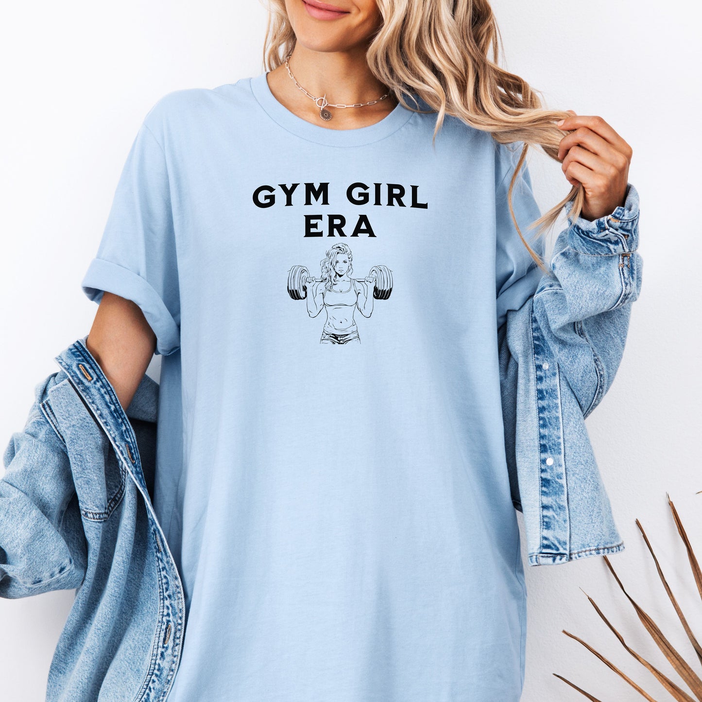 A Chambray light blue T-shirt with the bold black text “Gym Girl Era” above a line art illustration of a muscular woman lifting dumbbells. The fitness-inspired design celebrates strength, empowerment, and dedication to the gym lifestyle. Paired with denim shorts and a jean jacket, the look is casual yet powerful.