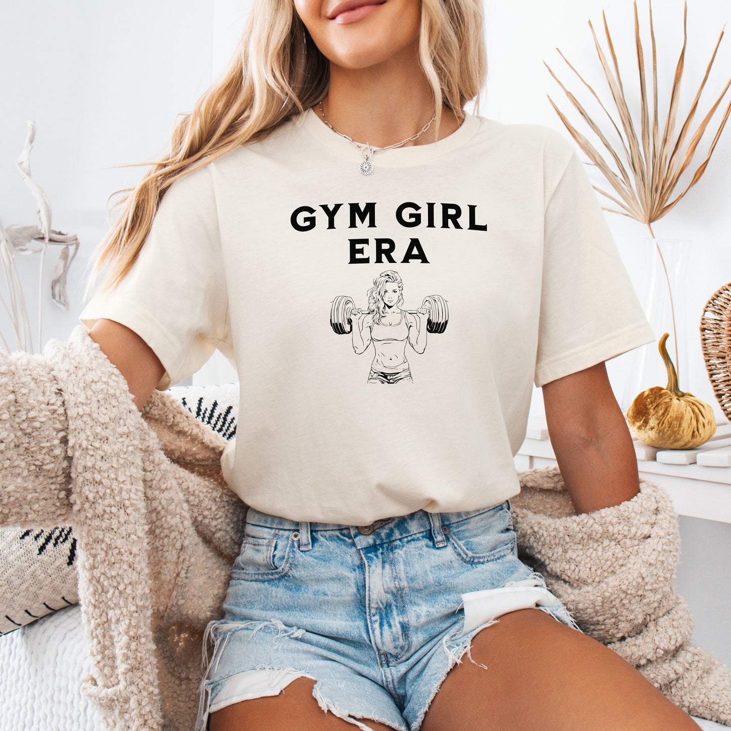 An ivory T-shirt with the bold black text “Gym Girl Era” above a line art illustration of a muscular woman lifting dumbbells. The fitness-inspired design celebrates strength, empowerment, and dedication to the gym lifestyle. Paired with denim shorts and a jean jacket, the look is casual yet powerful.