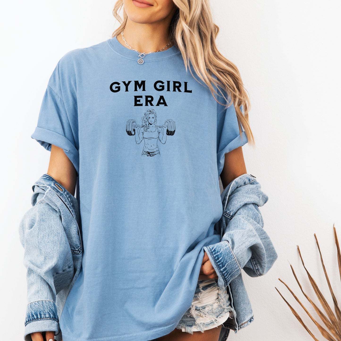 A washed denim T-shirt with the bold black text “Gym Girl Era” above a line art illustration of a muscular woman lifting dumbbells. The fitness-inspired design celebrates strength, empowerment, and dedication to the gym lifestyle. Paired with denim shorts and a jean jacket, the look is casual yet powerful.
