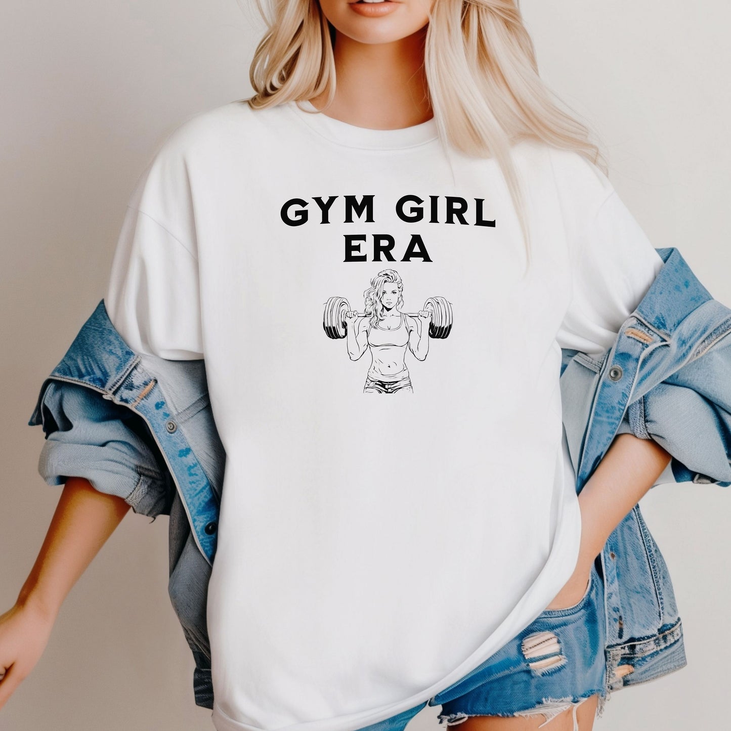 A classic white T-shirt with the bold black text “Gym Girl Era” above a line art illustration of a muscular woman lifting dumbbells. The fitness-inspired design celebrates strength, empowerment, and dedication to the gym lifestyle. Paired with denim shorts and a jean jacket, the look is casual yet powerful.