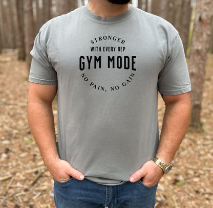 A person wearing a grey T-shirt with the bold black text “Stronger With Every Rep – Gym Mode – No Pain, No Gain.” The fitness-themed design features a curved text layout, perfect for workout enthusiasts and gym-goers who embrace strength training and motivation.