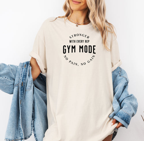 A person wearing an ivory T-shirt with the bold black text “Stronger With Every Rep – Gym Mode – No Pain, No Gain.” The fitness-themed design features a curved text layout, perfect for workout enthusiasts and gym-goers who embrace strength training and motivation.