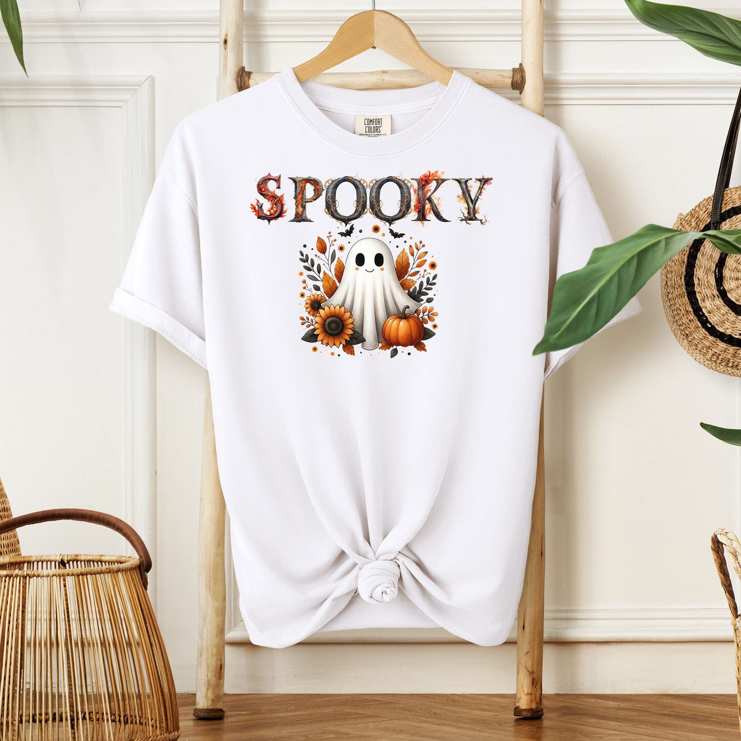 White T-shirt featuring a cute "Spooky" Halloween design with a smiling ghost surrounded by pumpkins, sunflowers, and autumn leaves. The word "Spooky" is styled with rustic wood-textured letters accented with fall foliage. Perfect for festive fall outfits, Halloween parties, or cozy seasonal wear.