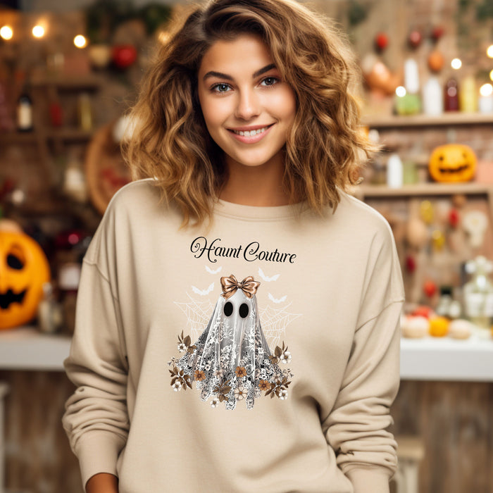 Sand Gildan sweatshirt featuring a stylish “Haunt Couture” design with a chic ghost draped in lace, wearing a metallic bow, surrounded by floral accents, spiderwebs, and flying bats. A glamorous and spooky Halloween look that blends fashion and festive charm—perfect for fall outings and haunted style.