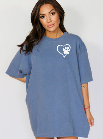 Person wearing a classic blue jean T-shirt featuring a simple white heart outline with a paw print inside on the upper left chest. The minimalist Heart Paw Print design symbolizes love for pets, making it a perfect shirt for dog and cat lovers.