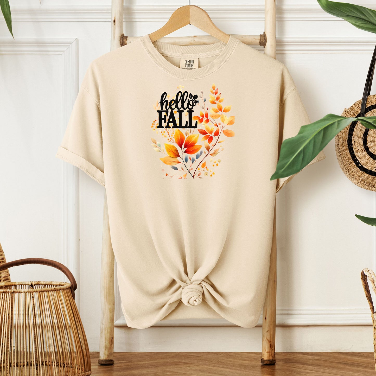 Ivory T-shirt featuring a vibrant "Hello Fall" design with hand-painted autumn leaves in orange, yellow, and soft grey blue tones. The bold black lettering is accented by delicate foliage, giving this seasonal tee a cozy, cheerful, and nature-inspired look perfect for fall fashion.