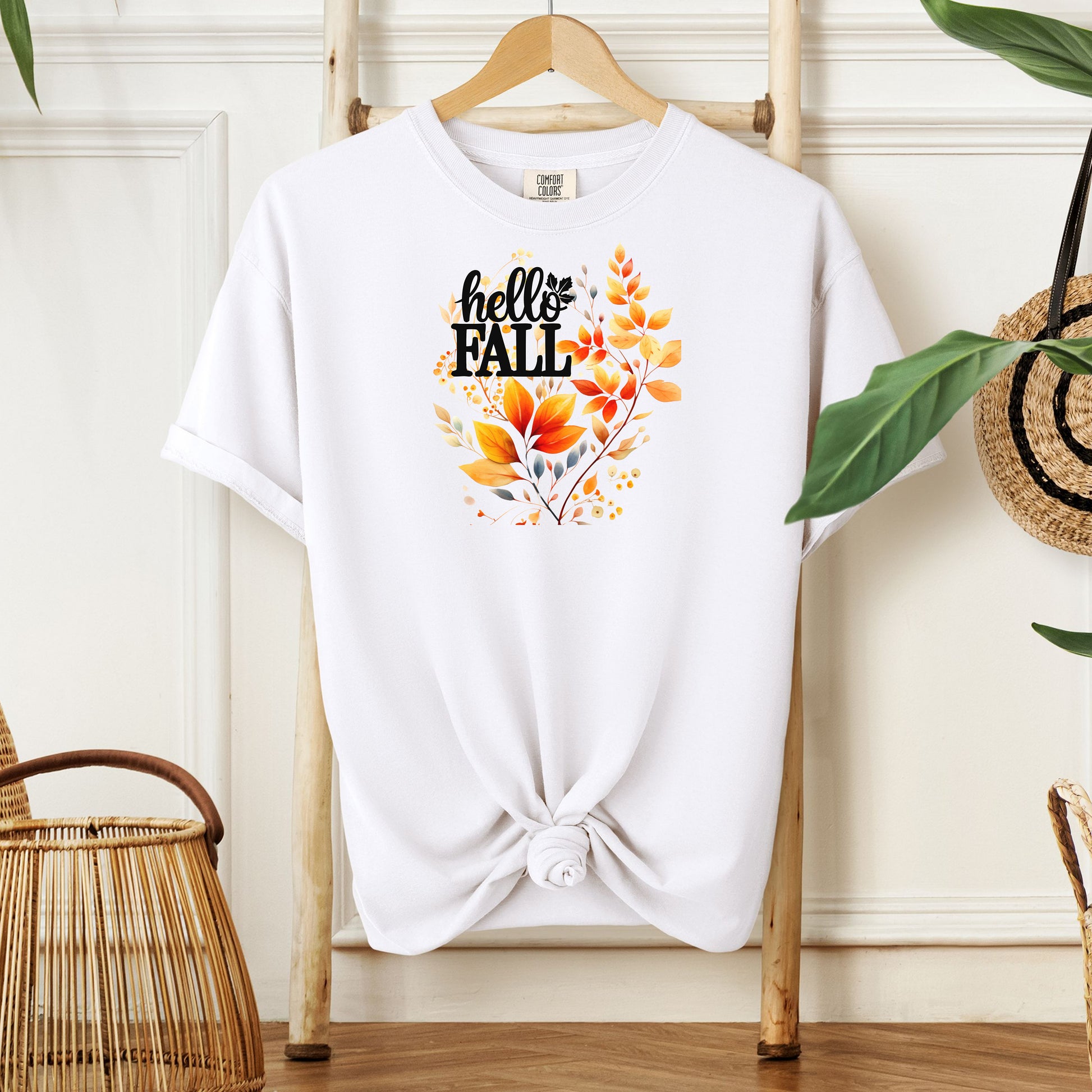 White T-shirt featuring a vibrant "Hello Fall" design with hand-painted autumn leaves in orange, yellow, and soft grey blue tones. The bold black lettering is accented by delicate foliage, giving this seasonal tee a cozy, cheerful, and nature-inspired look perfect for fall fashion.