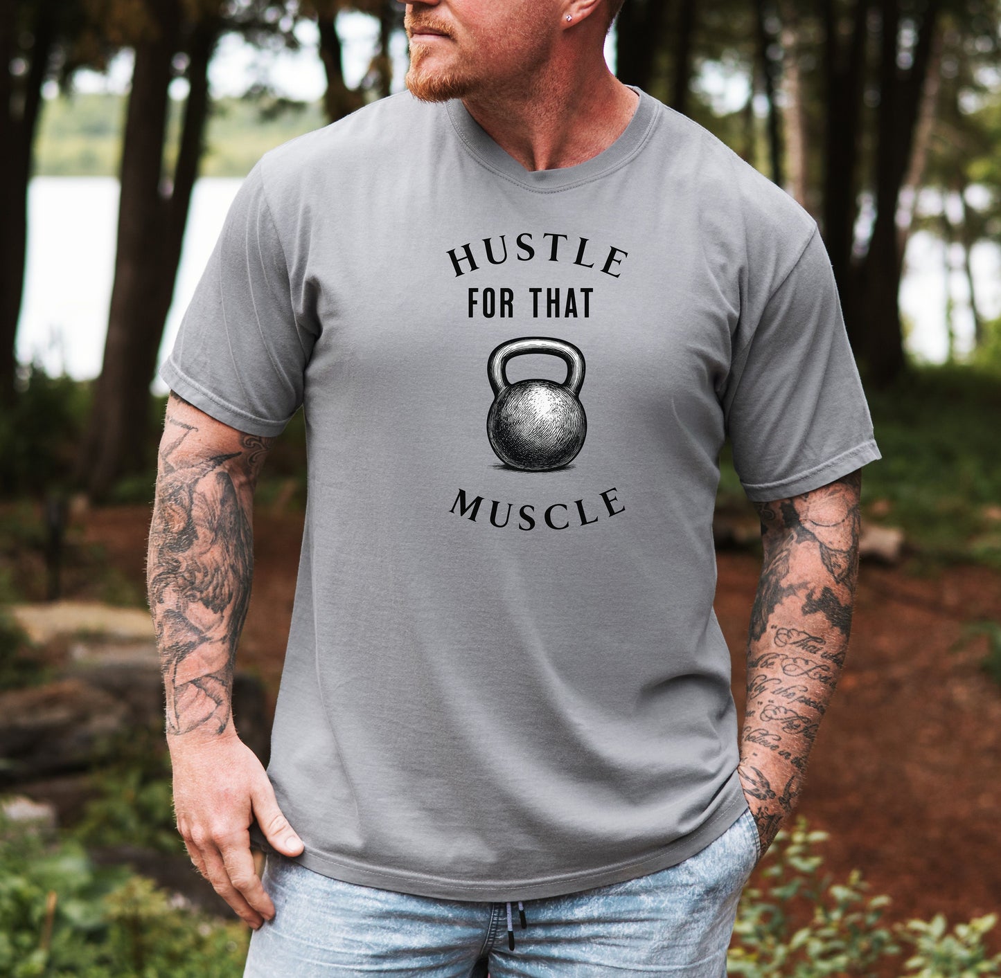 Person wearing a grey T-shirt with the phrase “Hustle For That Muscle” in bold black text, arched above and below a detailed kettlebell graphic. The design blends minimalist style with fitness motivation, perfect for gym-goers, weightlifters, and strength training enthusiasts who embrace hard work and dedication in their workout journey.