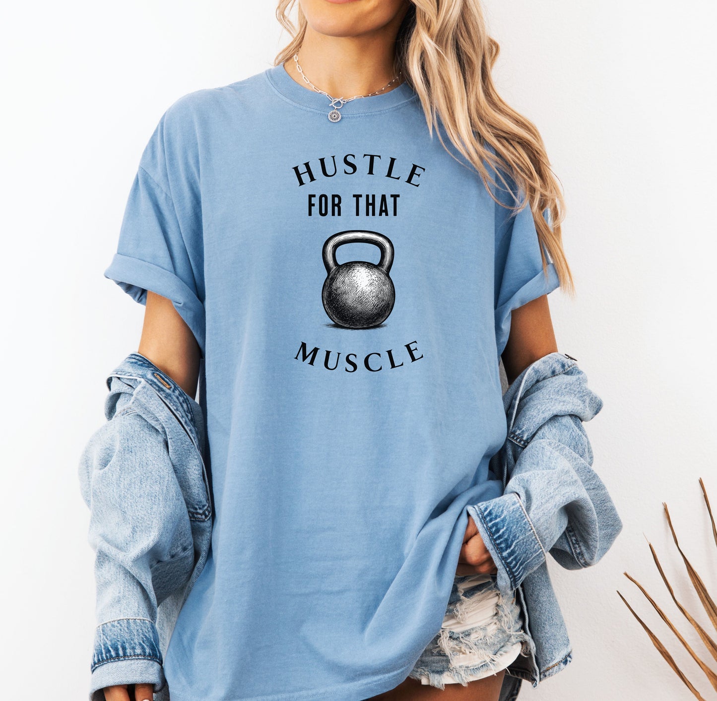 Person wearing a washed denim T-shirt with the phrase “Hustle For That Muscle” in bold black text, arched above and below a detailed kettlebell graphic. The design blends minimalist style with fitness motivation, perfect for gym-goers, weightlifters, and strength training enthusiasts who embrace hard work and dedication in their workout journey.