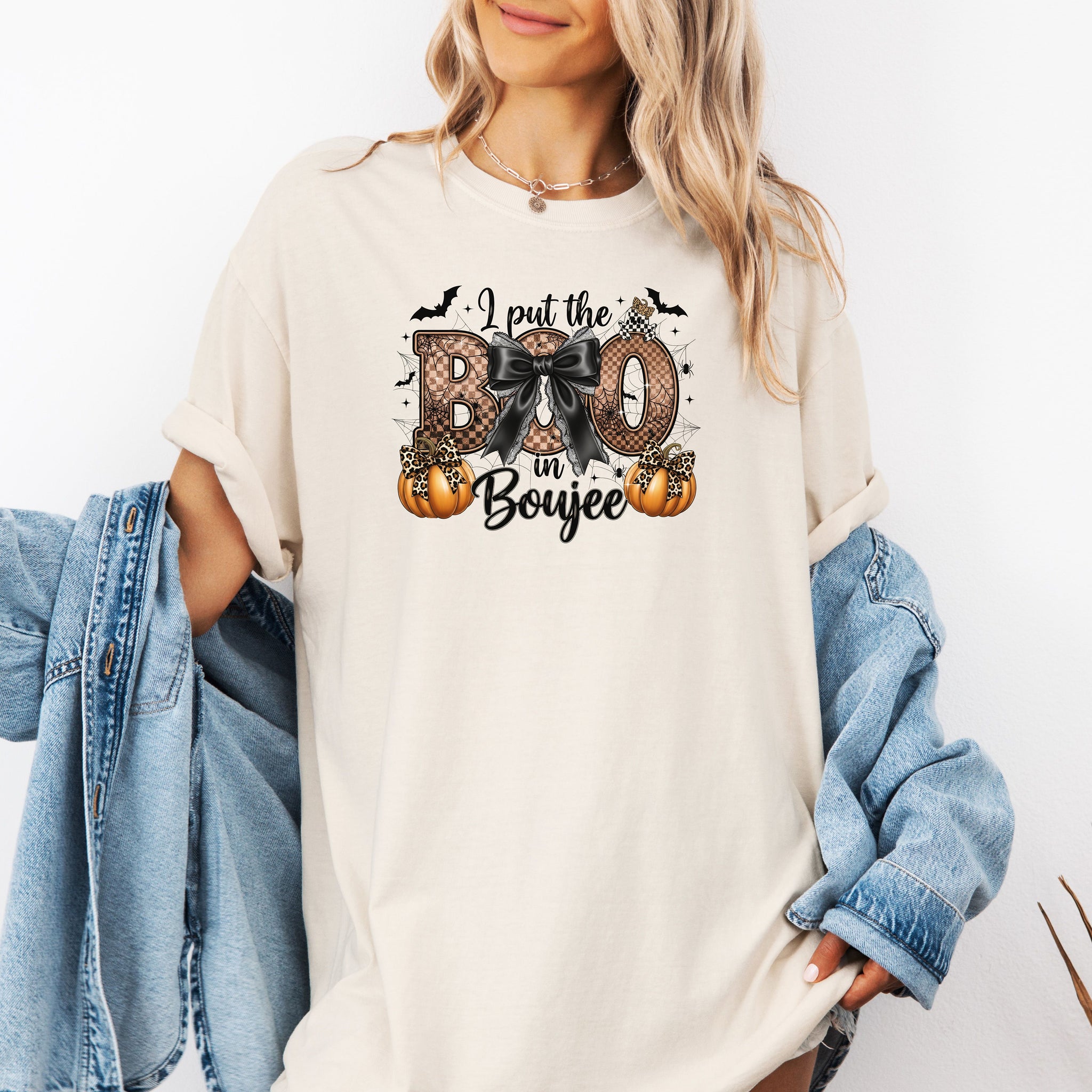 Person wearing an Ivory t-shirt with the phrase "I put the BOO in Boujee" in bold, Halloween-themed lettering. The design features a black bow, leopard print accents, bats, sparkling stars, and orange pumpkins, combining spooky and glamorous elements. Perfect for stylish Halloween lovers with a boujee flair.