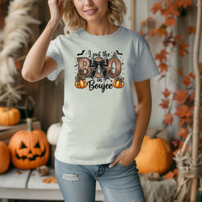 Person wearing a bay t-shirt with the phrase "I put the BOO in Boujee" in bold, Halloween-themed lettering. The design features a black bow, leopard print accents, bats, sparkling stars, and orange pumpkins, combining spooky and glamorous elements. Perfect for stylish Halloween lovers with a boujee flair.