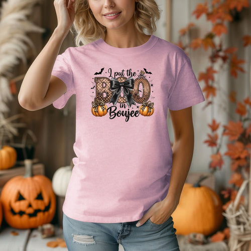 Person wearing a blossom soft pink t-shirt with the phrase "I put the BOO in Boujee" in bold, Halloween-themed lettering. The design features a black bow, leopard print accents, bats, sparkling stars, and orange pumpkins, combining spooky and glamorous elements. Perfect for stylish Halloween lovers with a boujee flair.