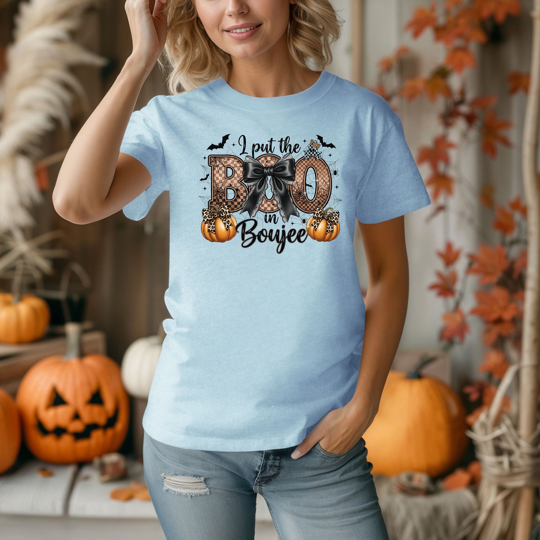 Person wearing a Chambray light blue t-shirt with the phrase "I put the BOO in Boujee" in bold, Halloween-themed lettering. The design features a black bow, leopard print accents, bats, sparkling stars, and orange pumpkins, combining spooky and glamorous elements. Perfect for stylish Halloween lovers with a boujee flair.