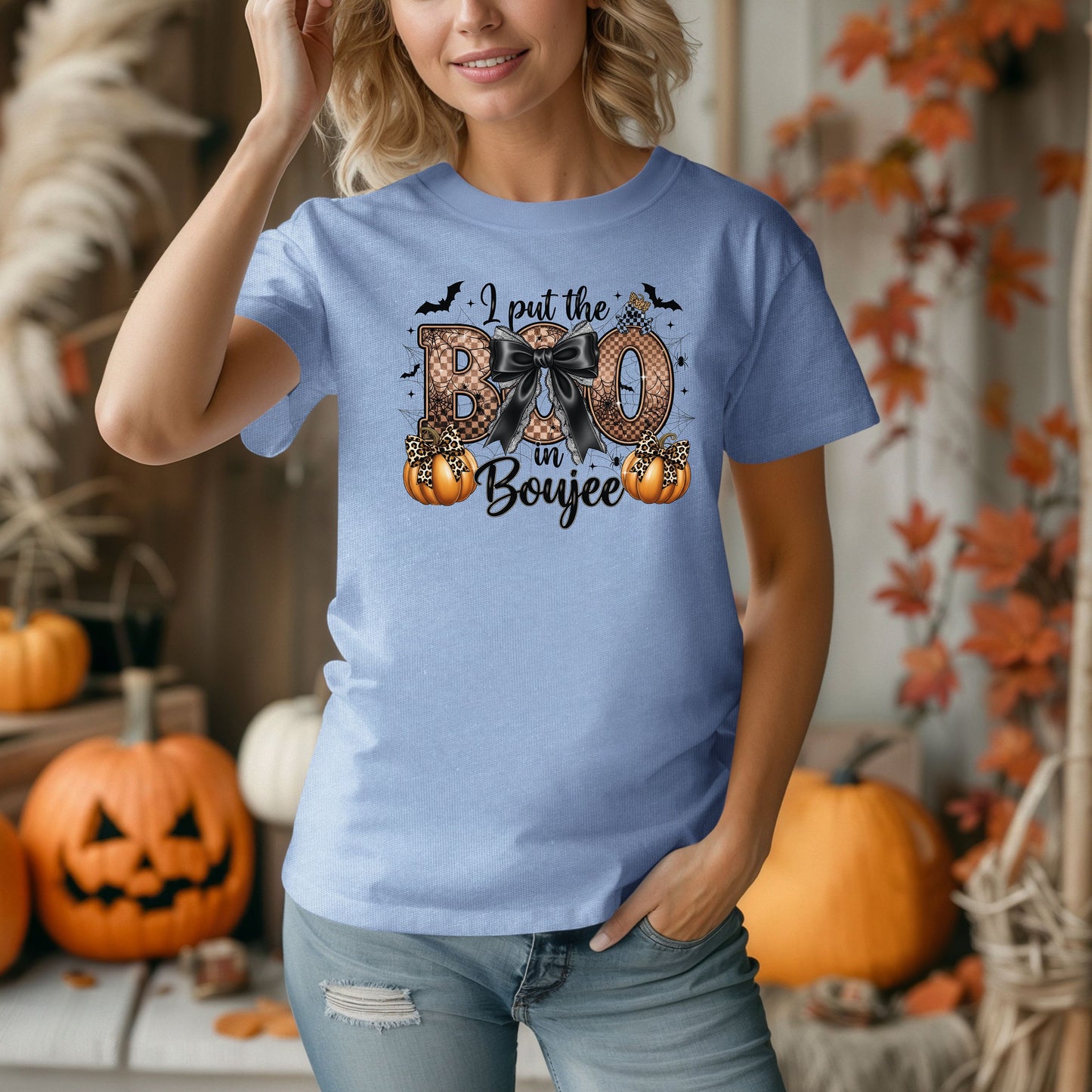 Person wearing a Washed Denim t-shirt with the phrase "I put the BOO in Boujee" in bold, Halloween-themed lettering. The design features a black bow, leopard print accents, bats, sparkling stars, and orange pumpkins, combining spooky and glamorous elements. Perfect for stylish Halloween lovers with a boujee flair.