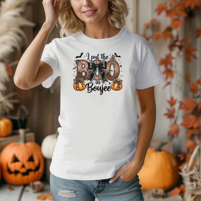Person wearing a white t-shirt with the phrase "I put the BOO in Boujee" in bold, Halloween-themed lettering. The design features a black bow, leopard print accents, bats, sparkling stars, and orange pumpkins, combining spooky and glamorous elements. Perfect for stylish Halloween lovers with a boujee flair.