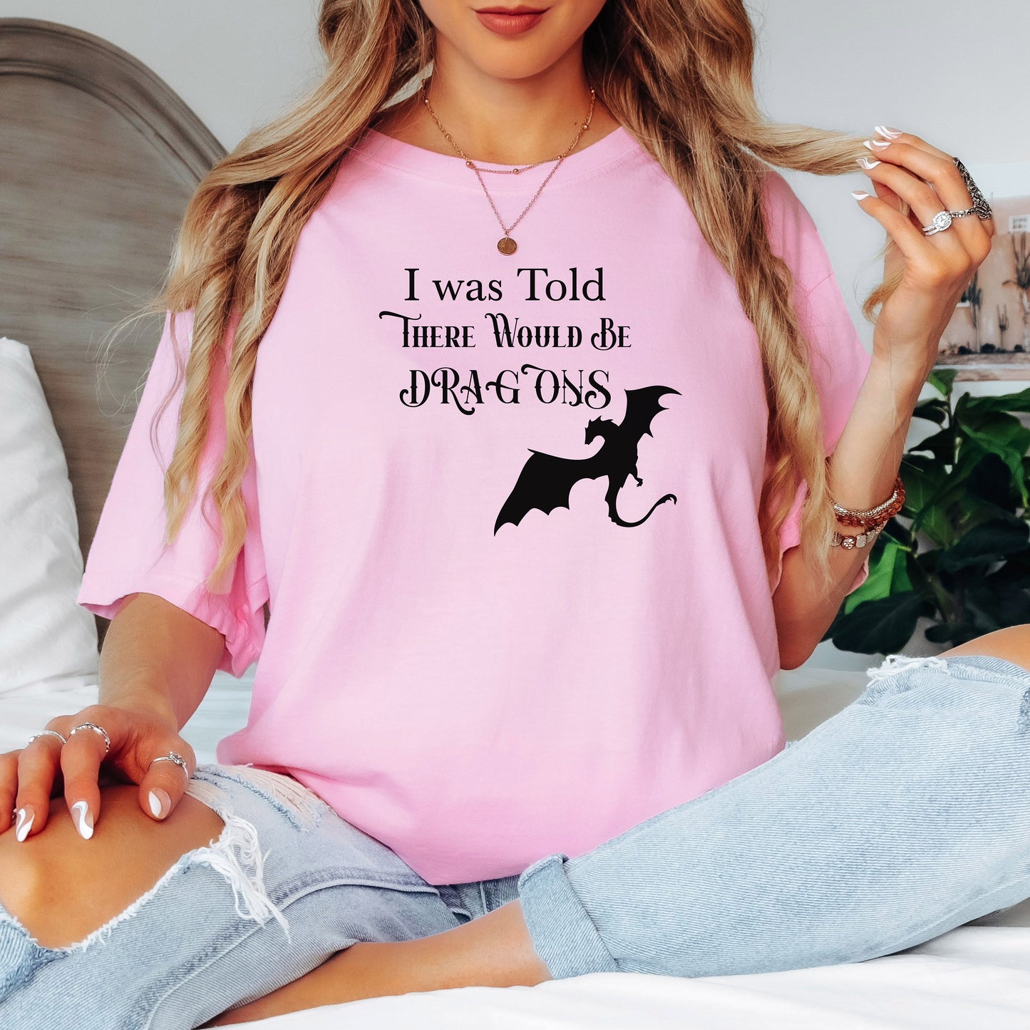 Person wearing a Blossom pink T-shirt with a Renaissance Faire-inspired design that reads “I Was Told There Would Be Dragons” in medieval-style fonts. The graphic features a bold silhouette of a flying dragon, perfect for fantasy lovers and fairgoers who enjoy whimsical and mythical-themed apparel.