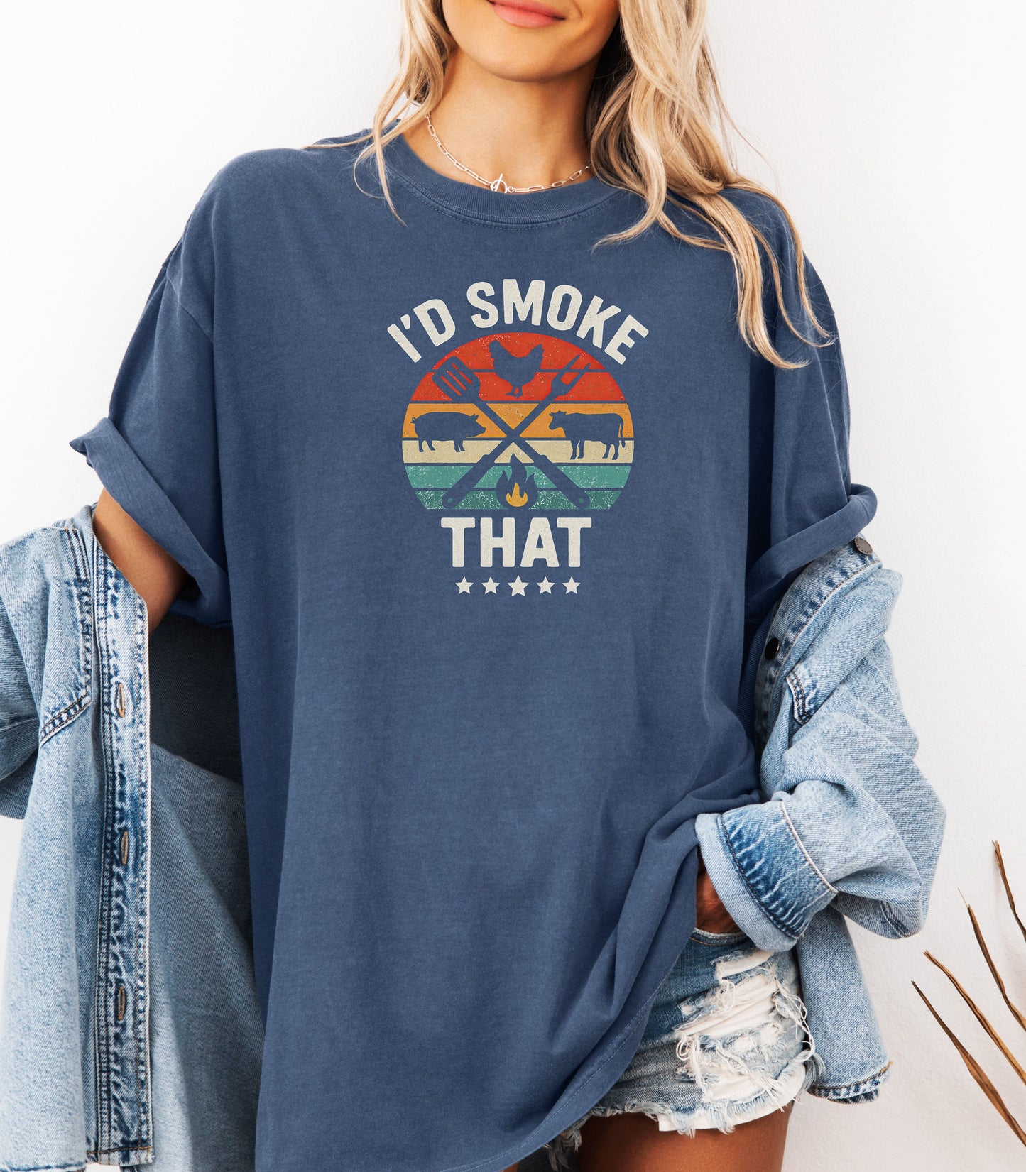 I’d Smoke That T-shirt featuring a retro sunset-style circle graphic with silhouettes of a chicken, pig, and cow, crossed grilling utensils, and a small flame. Text appears above and below the design, accented by five stars at the bottom. Printed on a denim unisex tee, this fun BBQ-themed shirt is perfect for grill masters, smoking enthusiasts, and meat lovers who enjoy cooking with humor.