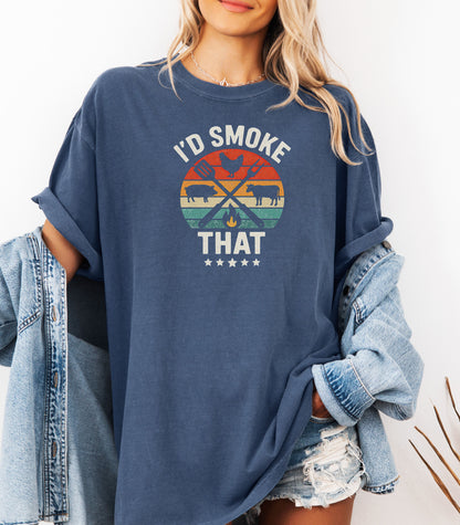 I’d Smoke That T-shirt featuring a retro sunset-style circle graphic with silhouettes of a chicken, pig, and cow, crossed grilling utensils, and a small flame. Text appears above and below the design, accented by five stars at the bottom. Printed on a denim unisex tee, this fun BBQ-themed shirt is perfect for grill masters, smoking enthusiasts, and meat lovers who enjoy cooking with humor.