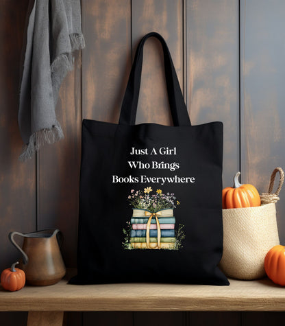 Black tote bag with 'Just A Girl Who Brings Books Everywhere' text and book illustration, surrounded by flowers.