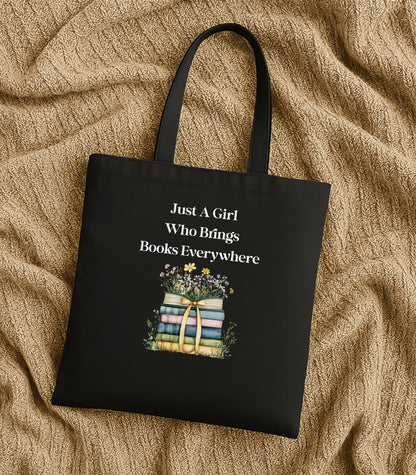 Black tote bag with 'Just A Girl Who Brings Books Everywhere' text and book illustration, surrounded by flowers.
