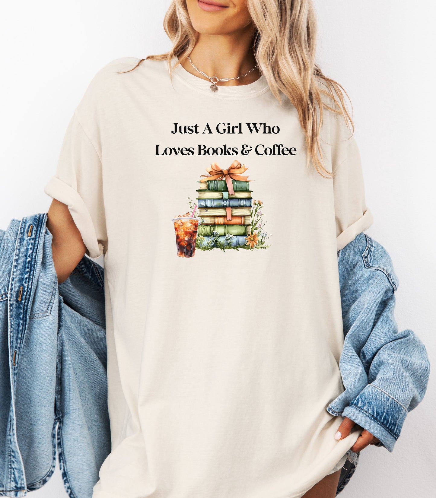 Woman wearing an Ivory t-shirt with the phrase "Just A Girl Who Loves Books & Coffee" printed above a charming stack of pastel-colored books tied with a ribbon, surrounded by fall foliage and a glass of iced coffee. A cozy and whimsical design perfect for bookworms and coffee lovers alike.