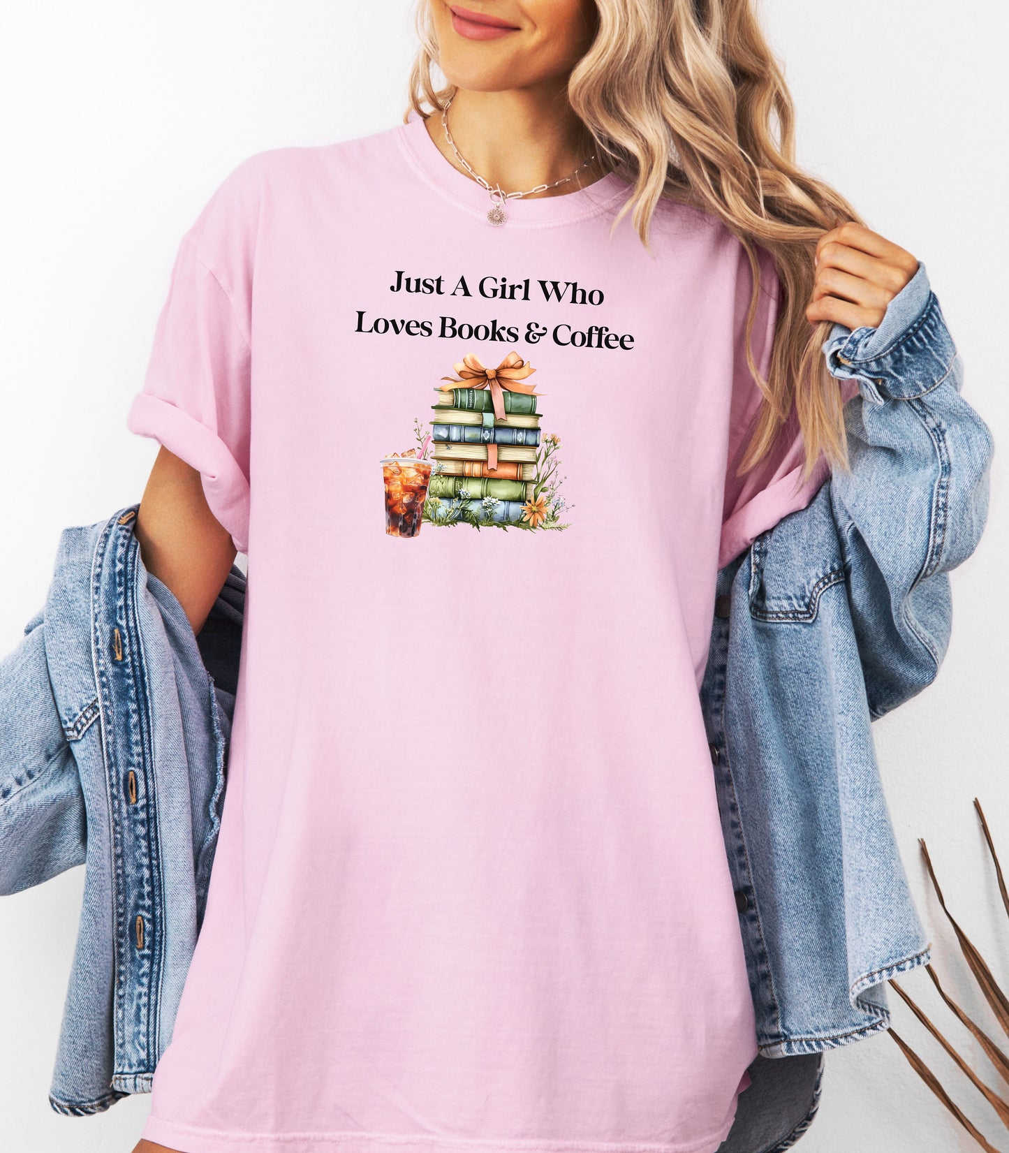 Woman wearing a blossom soft pink t-shirt with the phrase "Just A Girl Who Loves Books & Coffee" printed above a charming stack of pastel-colored books tied with a ribbon, surrounded by fall foliage and a glass of iced coffee. A cozy and whimsical design perfect for bookworms and coffee lovers alike.