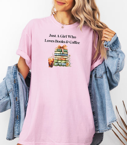 Woman wearing a blossom soft pink t-shirt with the phrase "Just A Girl Who Loves Books & Coffee" printed above a charming stack of pastel-colored books tied with a ribbon, surrounded by fall foliage and a glass of iced coffee. A cozy and whimsical design perfect for bookworms and coffee lovers alike.